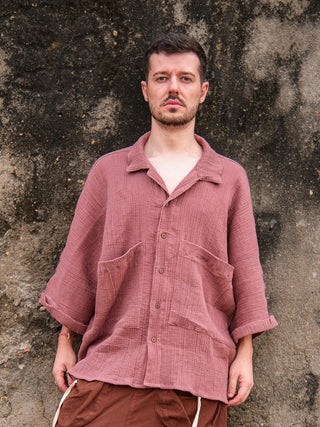 A man with short hair and a trimmed beard stands before a weathered wall, wearing the pink Yugma Baggy Shirt featuring large pockets and an elegant design, paired with brown pants. He looks neutrally at the camera.