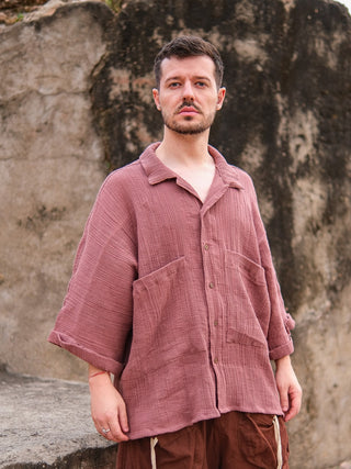 A man wearing the Yugma Baggy Shirt with large pockets and brown pants stands outdoors by a stone wall, looking at the camera.
