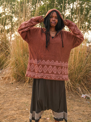 Standing amidst tall grass and trees, a person embodies the boho babe spirit in a Tribal Hoodie with geometric patterns, paired with a long skirt. They hold the hood while smiling.