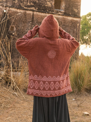 A person outdoors, facing away, is adjusting the hood of a rust-colored Tribal Hoodie with geometric patterns. In the background, tall grasses and a rustic building create a boho babe vibe.