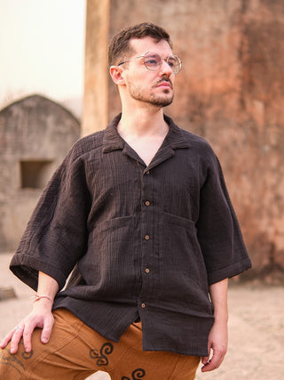 A man with short hair and glasses wears the Yugma Baggy Shirt, standing outdoors by a weathered stone wall in brown patterned pants, gazing thoughtfully to the side.