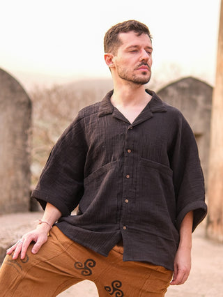 A man with short brown hair and a beard stands outdoors with his eyes closed and head tilted upward, wearing the Yugma Baggy Shirt made from premium quality fabric, paired with brown pants featuring black swirl designs.