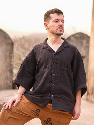 A man with short brown hair and a beard stands outdoors with his eyes closed and head tilted upward, wearing the Yugma Baggy Shirt made from premium quality fabric, paired with brown pants featuring black swirl designs.