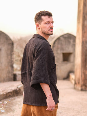A man with short hair and facial hair stands outdoors, wearing the Yugma Baggy Shirt and brown pants, looking over his shoulder against a backdrop of old stone structures and a soft, pale sky.