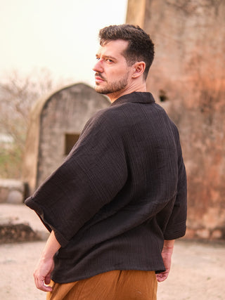 A man with short dark hair and a trimmed beard stands outdoors before an ancient stone structure, wearing brown pants and the Yugma Baggy Shirt made from premium quality fabric.
