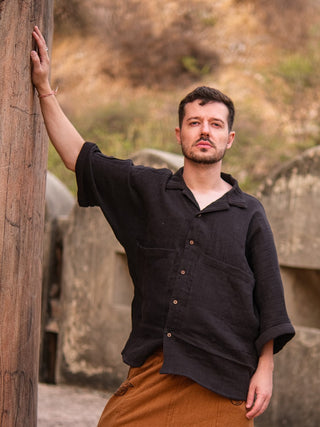 A man with short brown hair and a mustache wears a Yugma Baggy Shirt and brown pants, standing outdoors with one arm on a stone wall among weathered stone structures and dry vegetation.