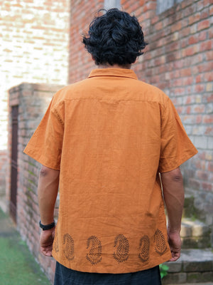A person with short curly black hair, wearing the elegant orange Udaya Shirt featuring decorative block print patterns near the hem, stands outside facing a brick wall.