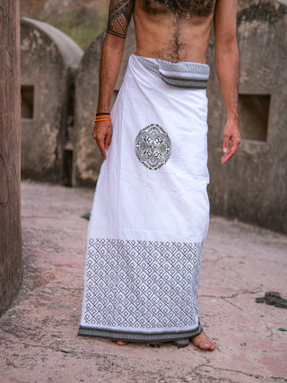 A man stands outdoors on a stone surface, wearing an Aastha Dhoti in white and green. His torso is bare, showing a tattoo on his upper arm and bracelets on his wrist.