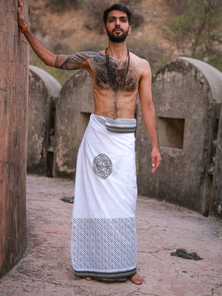 A shirtless man with arm tattoos stands against a rustic stone backdrop, wearing the Aastha Dhoti—crafted from cotton and featuring Rajasthani handblock print with a decorative emblem.