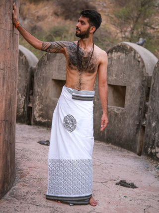A bearded, tattooed man stands outdoors in front of stone structures and dry vegetation, wearing the Aastha Dhoti—soft cotton in white with gray Rajasthani handblock print—and a necklace, one hand resting on a stone pillar.