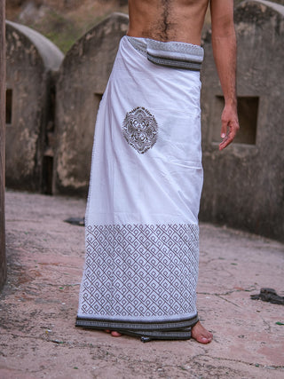 A man stands barefoot on a stone path, wearing the Aastha Dhoti—crafted from cotton with black geometric and circular Rajasthani handblock prints. His upper body is bare, and his arm rests at his side.