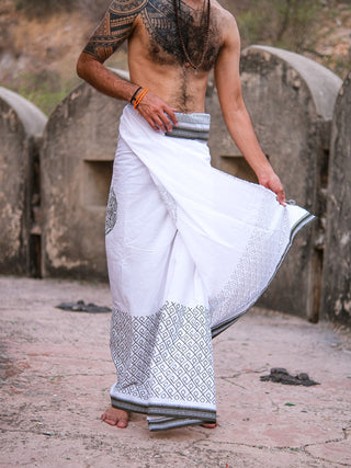 A man with tattoos and beaded necklaces stands barefoot on stone against an old wall, wearing the Aastha Dhoti—a traditional soft cotton white dhoti featuring Rajasthani handblock print borders.
