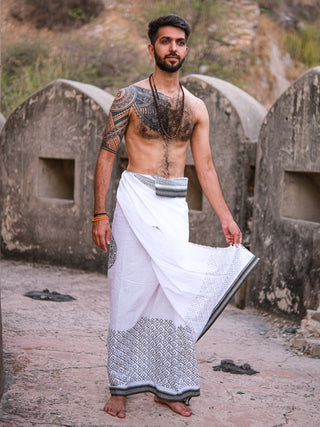 A bearded, tattooed man stands barefoot outdoors in front of stone walls and rocks, wearing the soft cotton Aastha Dhoti in traditional white and grey with necklaces.
