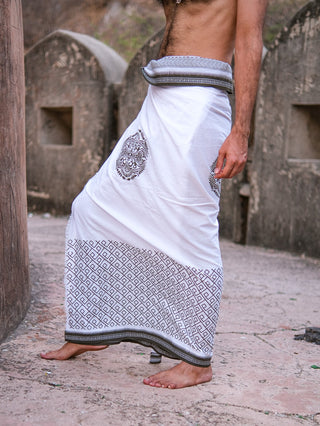 A barefoot man stands on stone, wearing the Aastha Dhoti—a white traditional garment made from soft cotton with black Rajasthani handblock print, draped around his waist down to his ankles. His upper body is not visible.