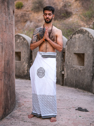 A man stands outdoors with palms pressed in prayer, wearing the Aastha Dhoti with Rajasthani handblock print borders and a beaded necklace, set against stone structures and dry vegetation.