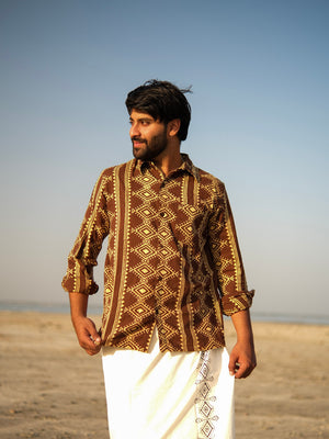 A man wearing the Varun Handblock Printed Cotton Shirt, featuring intricate Indian craftsmanship, and a white garment stands on a sandy beach under a clear blue sky, looking to his left with a slight smile.