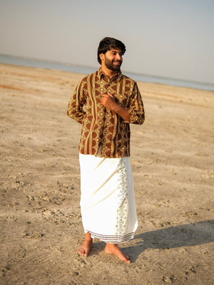 A man stands barefoot on sandy ground near the shore, wearing the Varun Handblock Printed Cotton Shirt that showcases Indian craftsmanship and a white traditional garment with a decorative border, looking to the side and smiling.