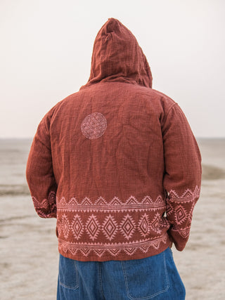 A person faces away from the camera in a rust-colored Tribal Hoodie, featuring geometric patterns on the shoulders and hem that blend seamlessly with the sandy backdrop, capturing a boho babe vibe.