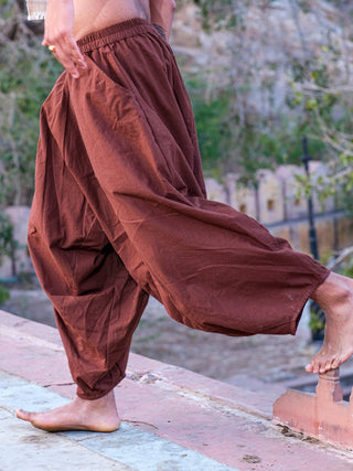 A barefoot person in brown Sirwal pants walks along an outdoor concrete ledge with a blurred, tree-filled background, showcasing a modern take on classic harem style.