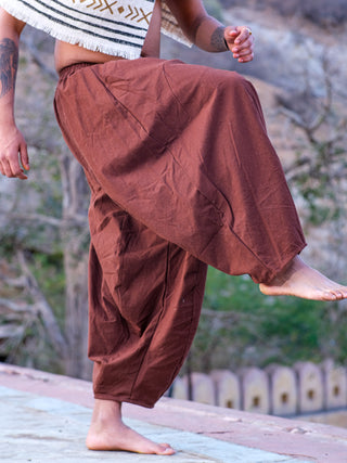 Wearing Sirwal pants, a person stands outdoors on stone, one bare foot raised. The loose brown pants and patterned shawl give a modern twist to traditional style, set against trees and a blurred background.