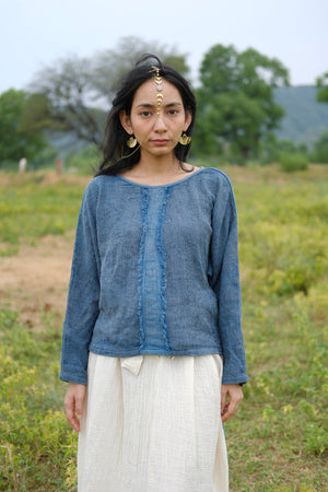 A woman stands outdoors in a grassy field, wearing the Bohemian Long-Sleeve Top and a white skirt, accessorized with gold earrings and a gold maang tikka on her forehead. Trees and hills in the background evoke the relaxed elegance of resort wear.