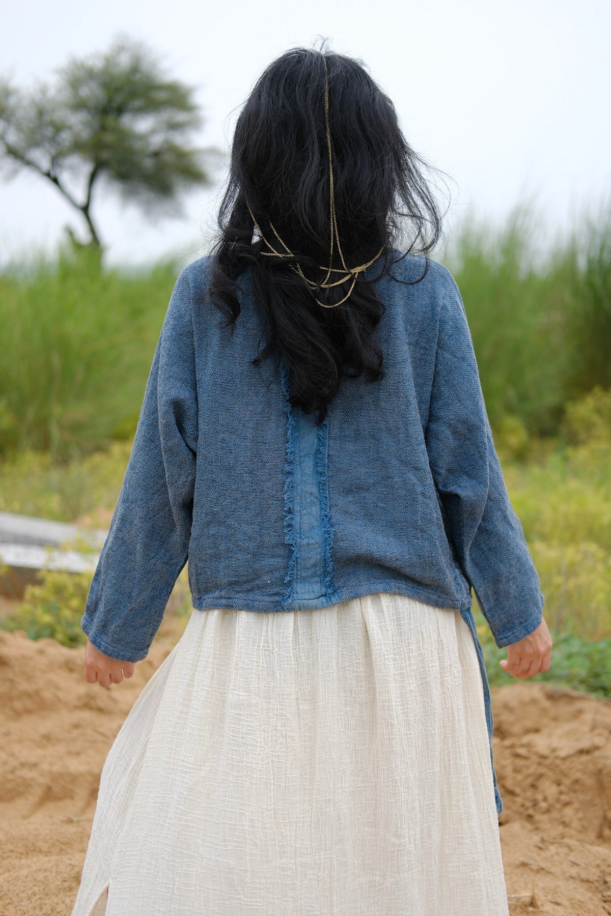 A person with long black hair, wearing the Bohemian Long-Sleeve Top and a white skirt, stands outdoors facing away, surrounded by greenery and a tree in the background. The outfit captures relaxed resort wear style—the Bohemian Long-Sleeve Top drapes effortlessly like a lightweight viscose knit, perfect for breezy days.