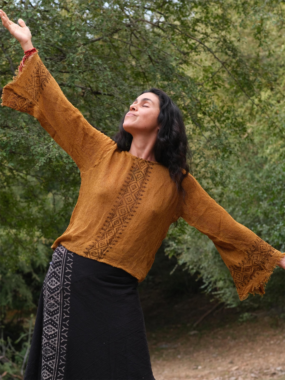 A person with long dark hair stands outdoors among lush green trees, eyes closed and arms raised joyfully, wearing the Kapisha Top paired with a black skirt.