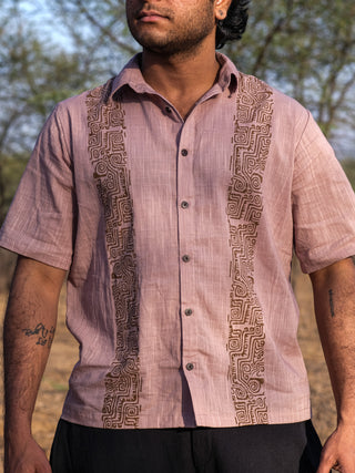 Someone wearing the Utopian shirt, featuring light pink fabric with detailed brown tribal-inspired geometric patterns, stands outdoors with blurred trees in the background.