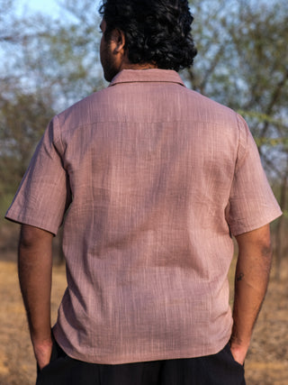 A man with curly hair stands outdoors, facing away, wearing a short-sleeved light brown Utopian shirt with a subtle tribal print and black pants. Trees and dry grass appear blurred in the background.