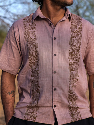 A person wearing a short-sleeved, Mose Utopian shirt with brown tribal designs along both sides of the buttons stands outdoors, their head slightly turned to the side.