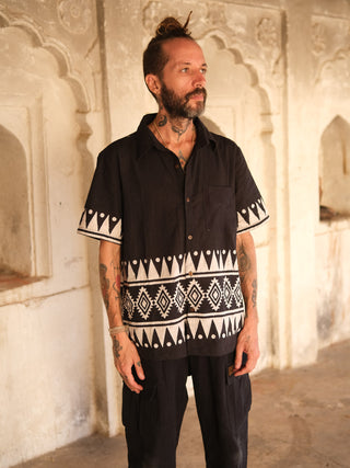 A tattooed individual stands before an ornate wall, wearing a Chakor Shirt with high-quality black and white patterns that match their black pants, adding to the scenes elegance with intricate arch designs in the background.