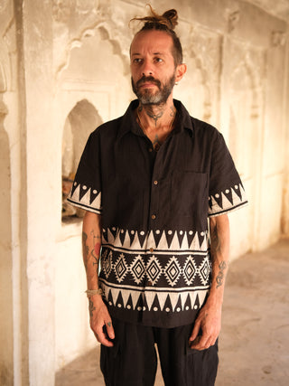 In a sunlit room with ornate white walls, a tattooed man with a bun wears the Chakor Shirt, distinguished by its white geometric pattern made from premium materials, paired seamlessly with black pants as he gazes slightly to the side.