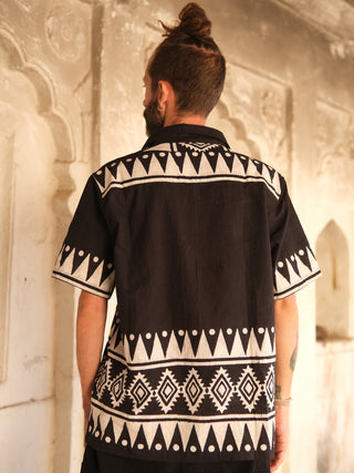 A man with a bun hairstyle stands back to the camera in a Chakor Shirt featuring white geometric patterns. The high-quality black fabric contrasts elegantly with the ornate, light stone architecture behind him.