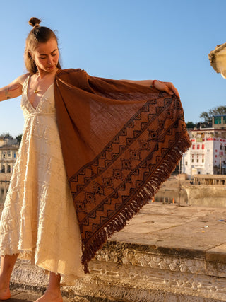 Standing on a stone ledge overlooking historic buildings and a clear blue sky, someone in a white dress holds the intricately patterned Neelkanth Shawl.