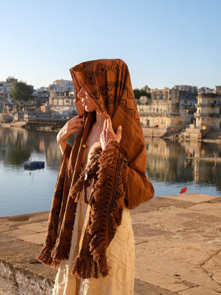 By the riverside, someone holds a brown Neelkanth Shawl close, its bohemian prints creating a serene scene with a small boat drifting under a clear blue sky and buildings in the backdrop.