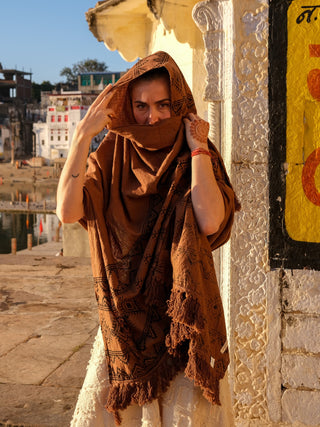 A person by the river, partially covering their face with a Neelkanth shawl in Rajasthani hand block print, showcases henna-adorned hands amidst a sunny backdrop of historic buildings and water.