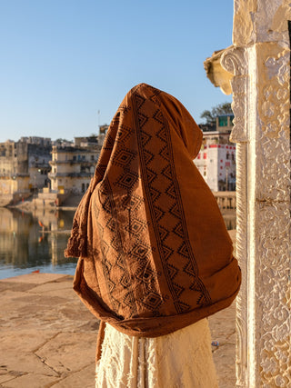 Wearing a Neelkanth Shawl, which evokes the charm of Rajasthani hand block print, a person stands by a tranquil lake. In the background, intricate carvings and historic buildings add to the serene scene beneath a clear sky.