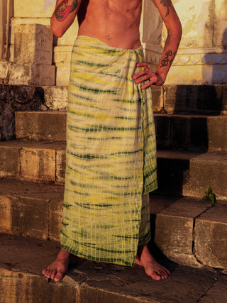 A person stands barefoot on stone steps, wrapped in a vibrant Leo Dhoti, sunlight illuminating their tattoos and the textured wall behind them.