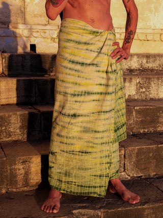 A barefoot person stands on stone steps, wearing the Leo Dhoti sarong in tie-dyed yellow and green around their waist, with a bare upper body, one hand resting on the sarong and tattoos visible on their arms.