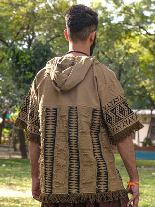 Facing away outdoors, a person wears the Sinai Hoodie, showcasing geometric patterns with fringed edges. This bohemian jacket mixes textures and colors, mainly brown and tan, against a backdrop of lush green trees.
