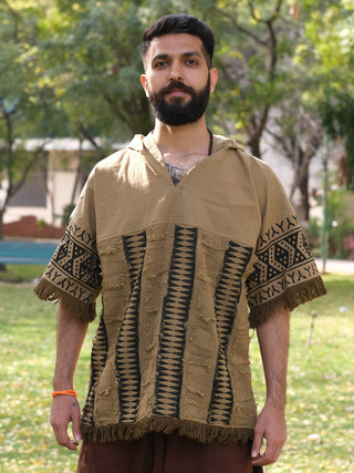 Outdoors in a park, a bearded man wears the Sinai Hoodie, a tan poncho-style garment with intricate bohemian patterns and fringe details. The greenery of trees and grass enhances the Rajasthani handblockprint artistry of his attire.