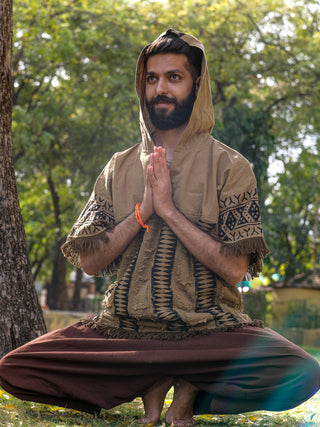 A bearded man sits cross-legged outdoors, clad in the Sinai Hoodie featuring Rajasthani handblock print. His hands are in a prayer position by his chest. In this bohemian park setting, hes surrounded by trees and grass as sunlight filters through the leaves.