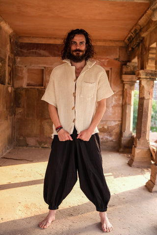 A barefoot man with long curly hair and a beard stands in a sunlit stone corridor, wearing the Amogh Shirt and loose black pants for subtle boho flair as he looks directly at the camera.