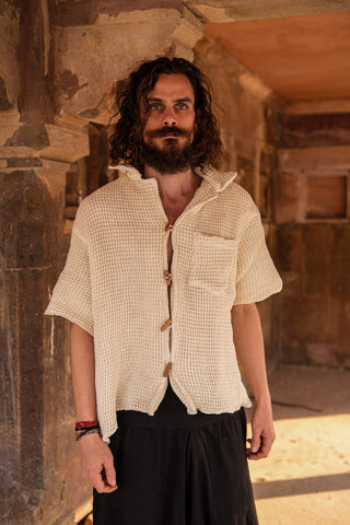 A man with long curly hair and a beard stands in a sunlit stone corridor, wearing the eco-friendly Amogh Shirt, featuring wooden buttons. He pairs it with a dark skirt and gazes at the camera, channeling subtle boho flair.