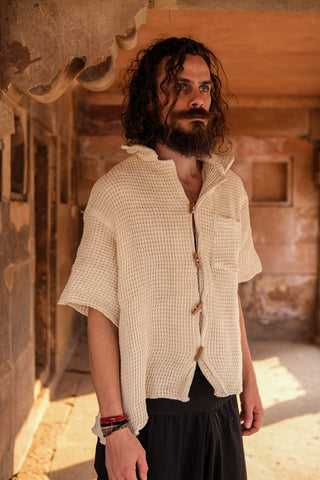 A curly-haired, bearded man stands in a sunlit stone corridor wearing the eco-friendly Amogh Shirt with wooden buttons and a pocket, paired with dark pants. He gazes to the side, exuding effortless boho style.