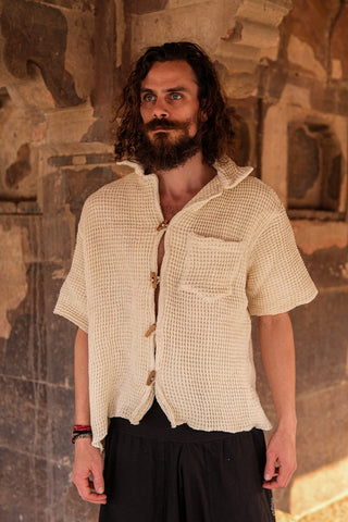 A man with curly hair and a beard stands by a stone wall, wearing the eco-friendly, cream-colored Amogh Shirt with short sleeves and textured fabric, paired with a black skirt for a stylish boho look.