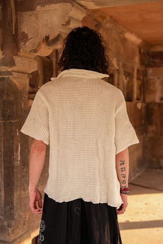 A person with curly hair faces away, wearing a loose, textured cream Amogh Shirt. Black pants and right forearm tattoos are visible, with stone architecture in the background.