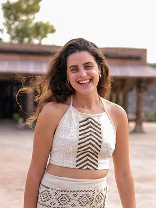 A woman stands outdoors, showcasing the Mathu Crop Top in a boho style paired with a patterned skirt. Her long, wavy hair flows gracefully against a rustic backdrop of trees and an old building.
