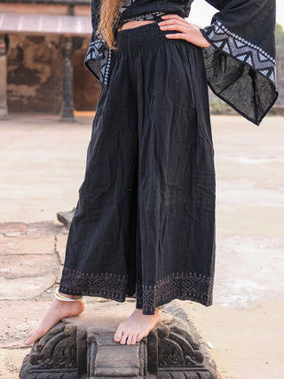 A person models boho-chic black Medusa Palazzo Pants with a matching top, barefoot on an ancient stone structure, hand on hip and one foot raised. Weathered stone architecture forms the backdrop.