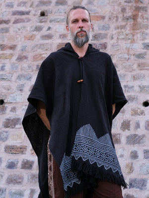 A bearded man stands against a stone wall, wearing a black hooded poncho adorned with light blue geometric patterns along the hem. Draped over his shoulders is the black Swing Shawl, complementing his outfit perfectly. With his hands tucked into his pockets, he gazes calmly at the camera.